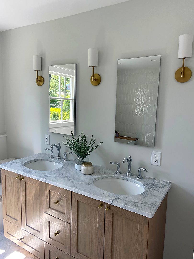 Oak double vanity with a marble top, brass wall sconces, and tall mirrors