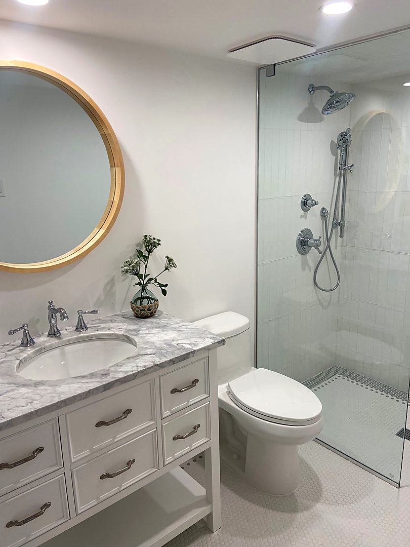 Bathroom with a marble-top white vanity, a round wood mirror, and a frameless glass shower