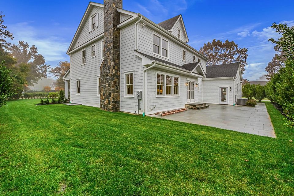 Rear of a white custom farmhouse with a stone chimney, a stone patio, and a green lawn