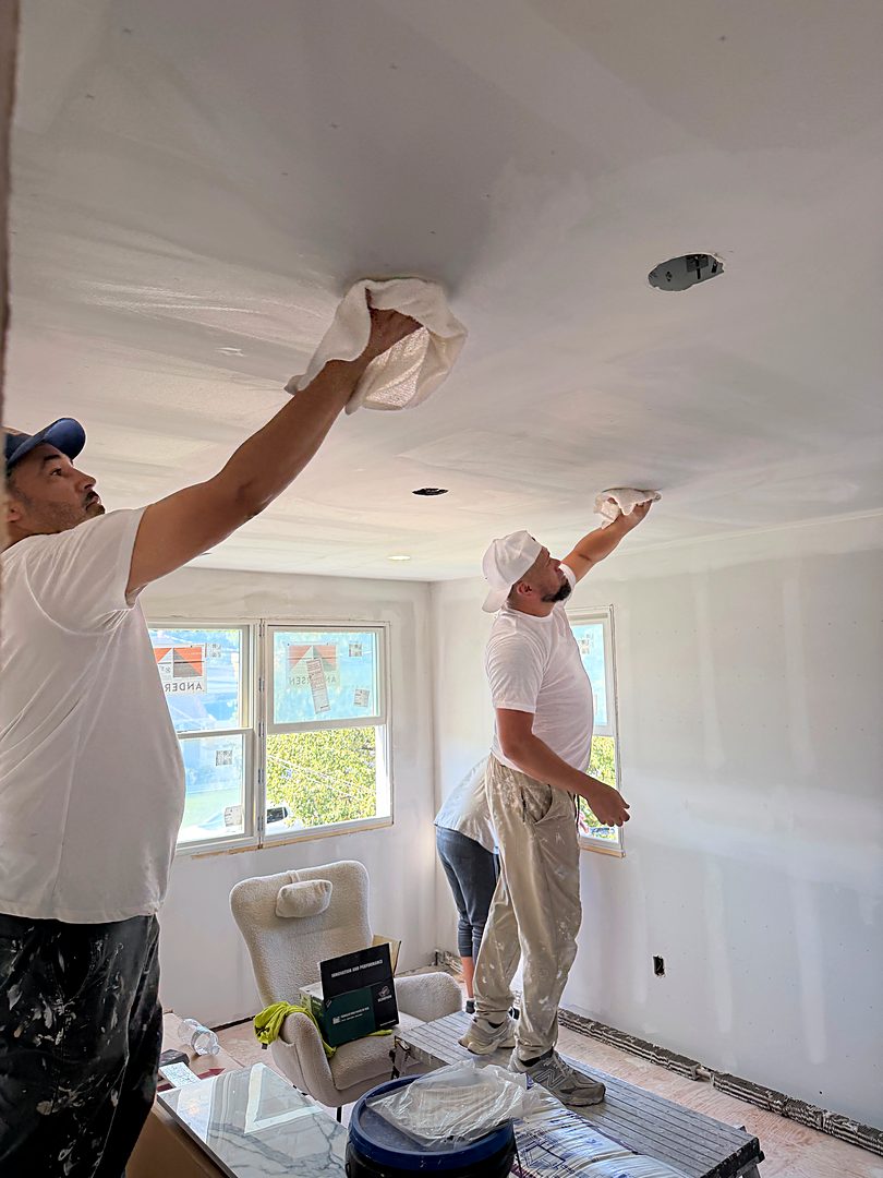 Two finishers skim coating a new drywall ceiling in a bedroom