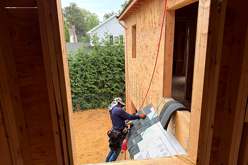 Crew member installing shingles on a lower roof, seen through a framed window opening