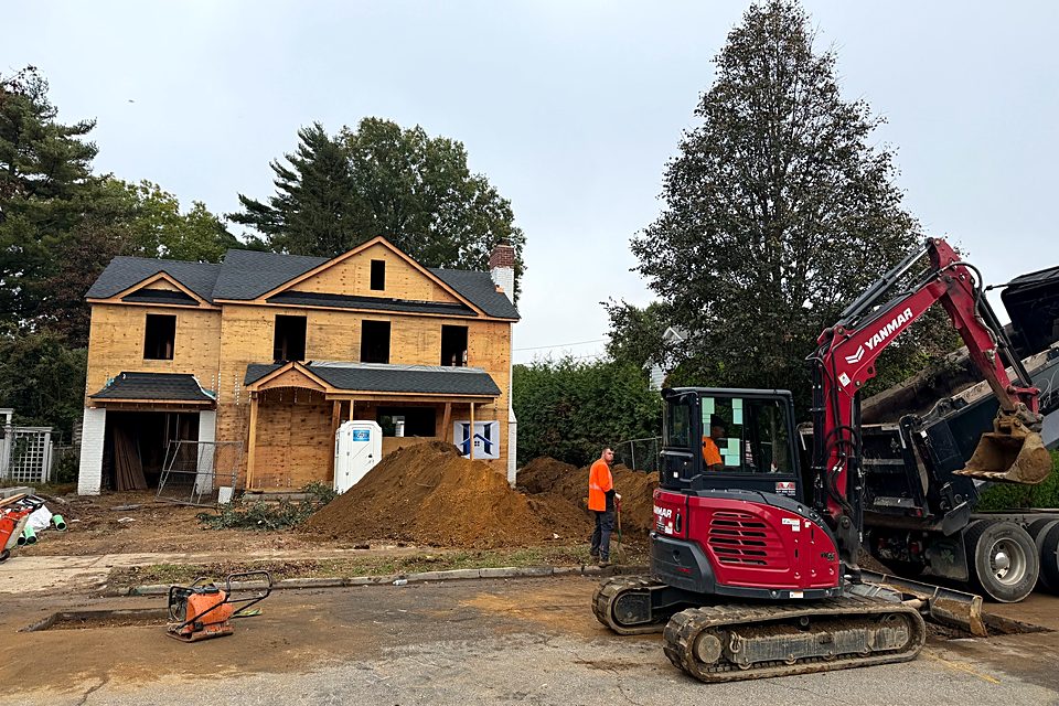 Excavator and crew at the street in front of the sheathed custom home, with dirt piled at the curb