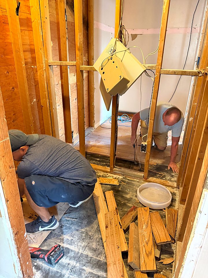 Two crew members working on the floor framing and drain in the opened bathroom